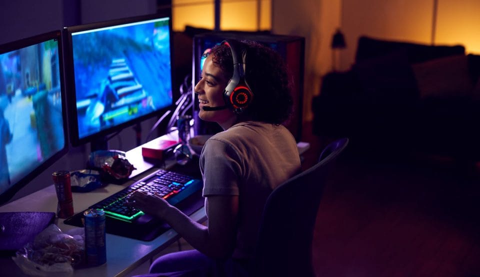 Teenage Girl Wearing Headset Gaming At Home Using Dual Computer Screens